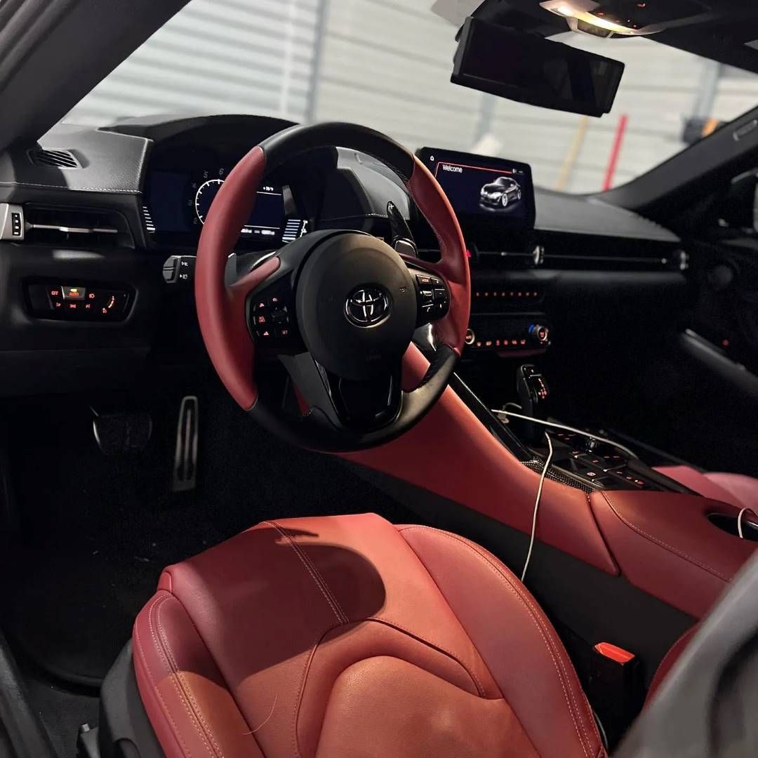 A close-up view of a Toyota Supra's luxurious red and black leather interior, highlighting the steering wheel and dashboard.
