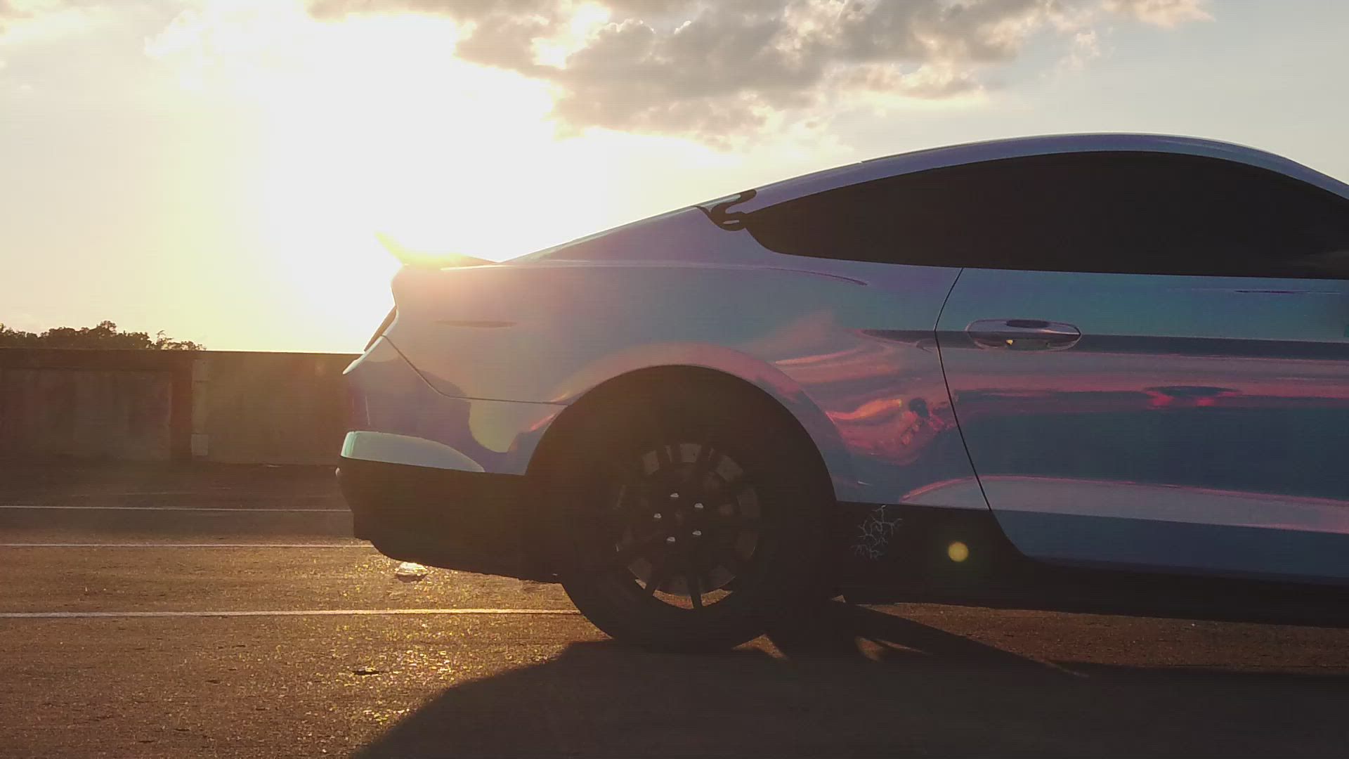 A sleek, iridescent car parked on a road at sunset, with the sun casting a warm glow and long shadows.