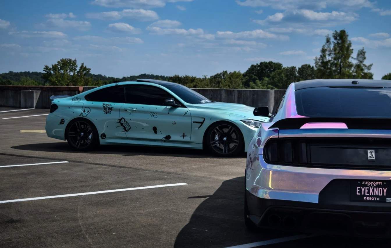 Two uniquely modified cars, one mint green with graffiti and one iridescent silver, parked side by side in an open lot under a partly cloudy sky.
