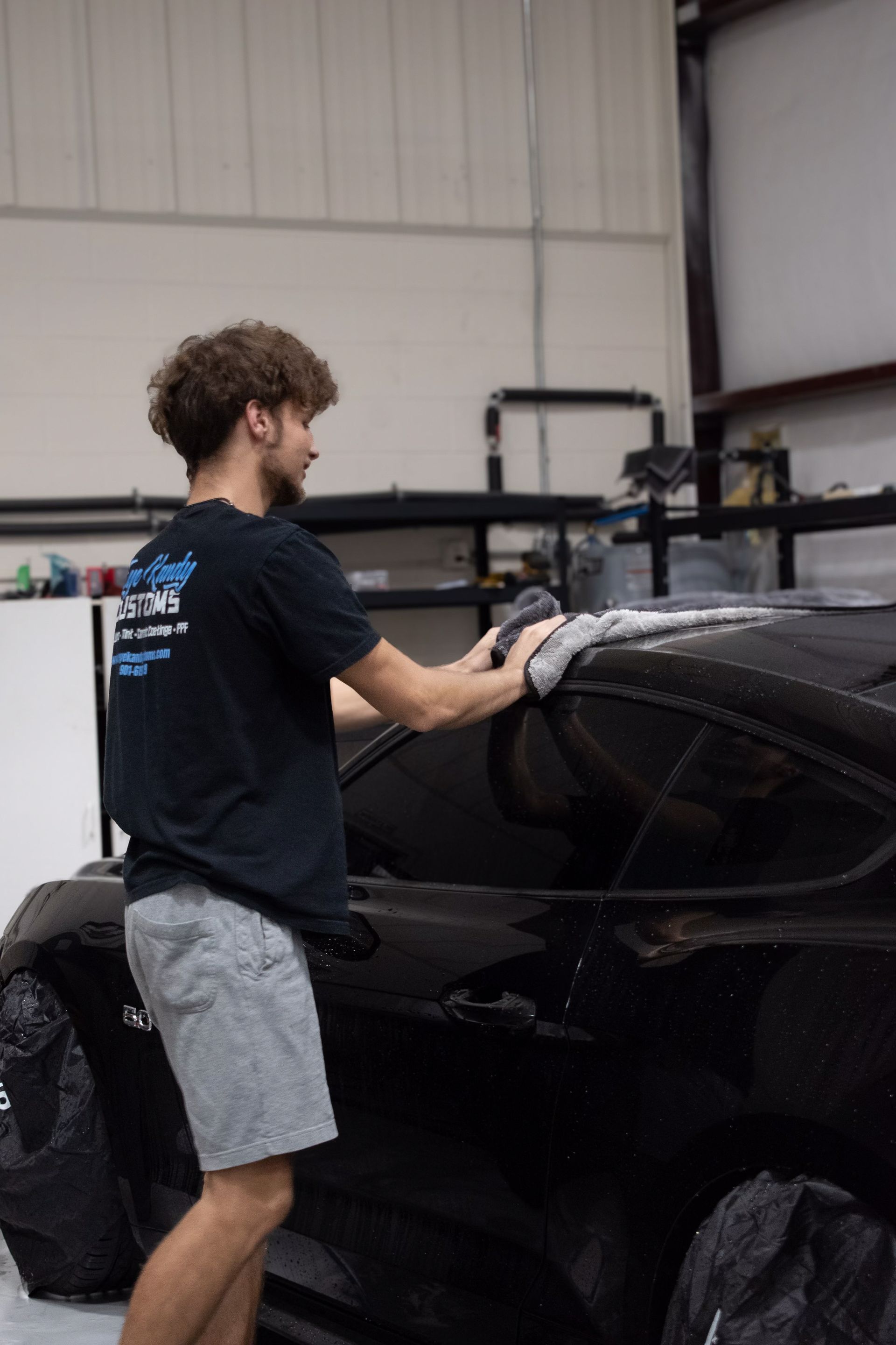 A man in a work shirt meticulously wipes down the hood of a black car with a microfiber cloth inside a garage.