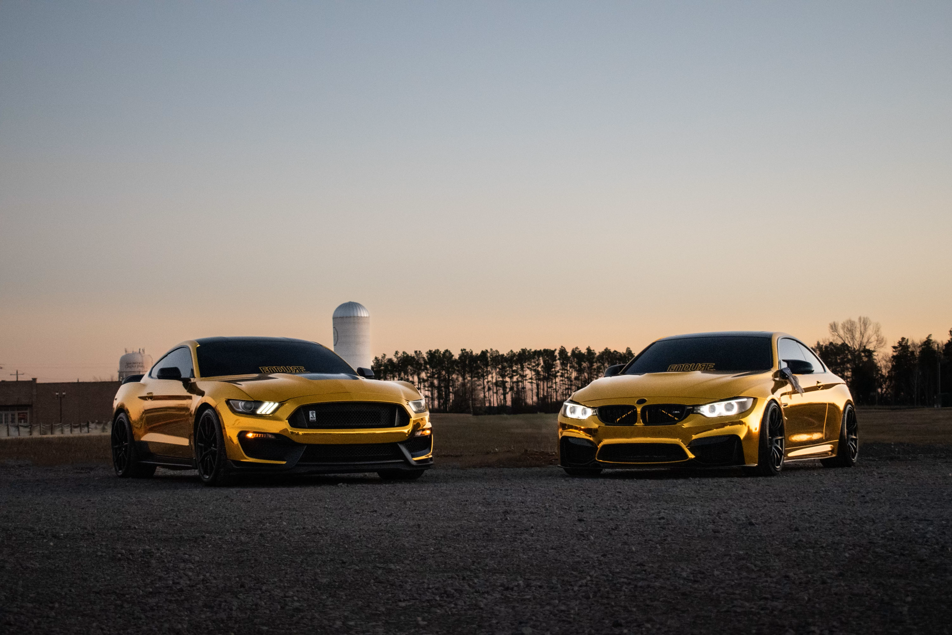 Two gold-painted sports cars face forward on a gravel lot at sunset, with silos and trees in the background.