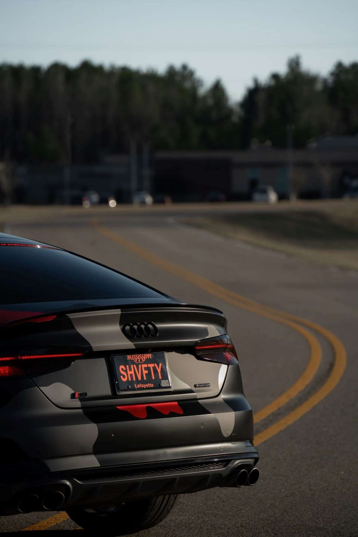 The rear view of a camouflaged Audi sedan with a custom license plate, parked on a curving road with trees in the background.