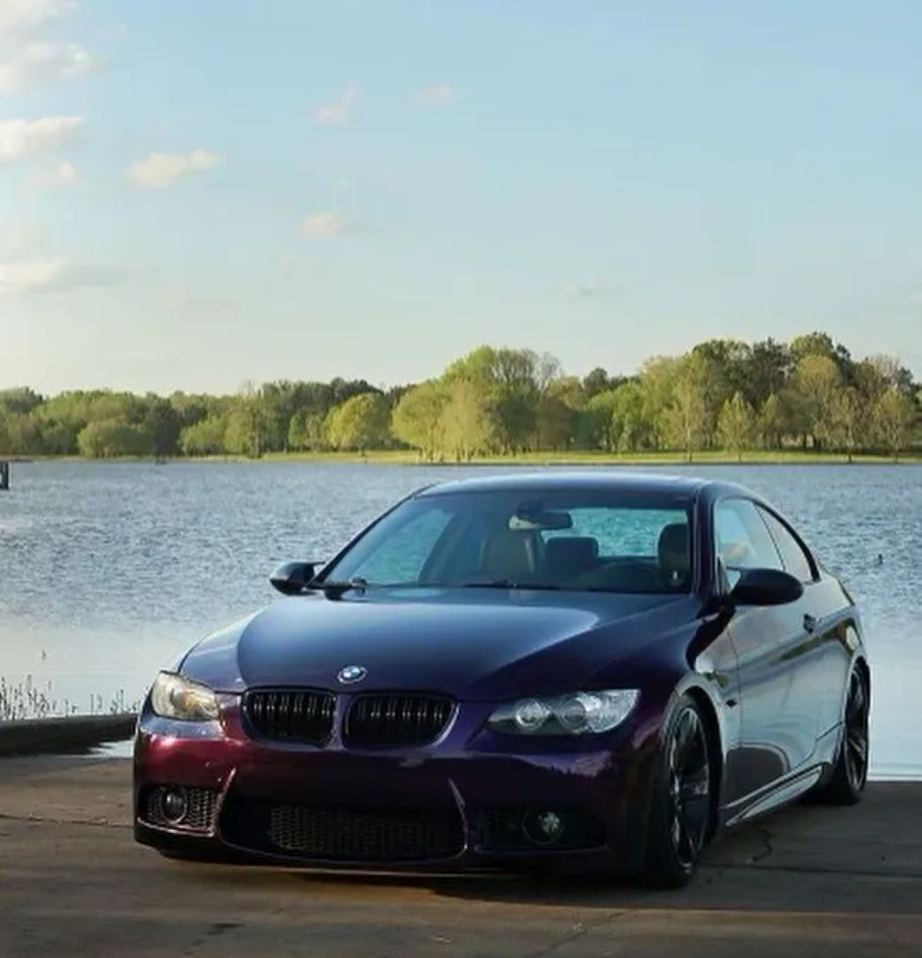 A glossy purple BMW coupe parked by a serene lake with trees in the background.